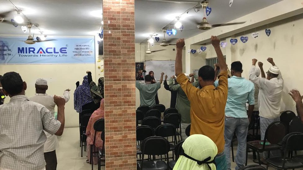 একজন নারী কর্মী মাইক ব্যবহার করে বেশ কয়েকজন বয়স্ক নারী-পুরুষকে ব্যায়াম করাচ্ছেন একজন নারী কর্মী মাইক ব্যবহার করে বেশ কয়েকজন বয়স্ক নারী-পুরুষকে ব্যায়াম করাচ্ছেন